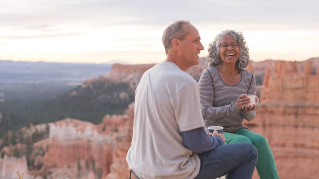 An older couple take a break from hiking to enjoy coffee on a Utah outlook overlooking a canyon.They are sitting on camp stools close to the edge. They are both holding coffee mugs and she is smiling at the camera.