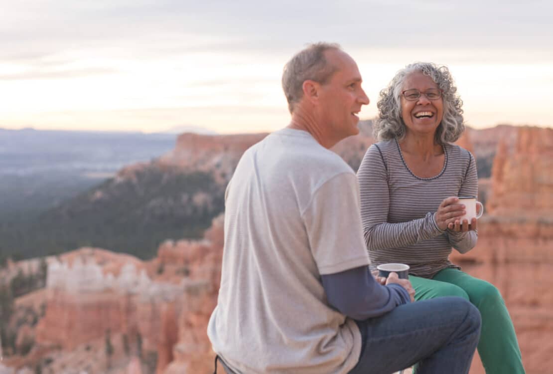 An older couple take a break from hiking to enjoy coffee on a Utah outlook overlooking a canyon.They are sitting on camp stools close to the edge. They are both holding coffee mugs and she is smiling at the camera.