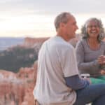 An older couple take a break from hiking to enjoy coffee on a Utah outlook overlooking a canyon.They are sitting on camp stools close to the edge. They are both holding coffee mugs and she is smiling at the camera.