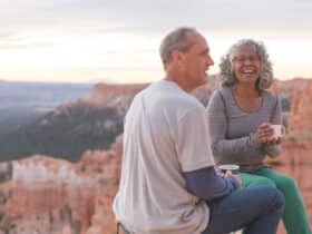 An older couple take a break from hiking to enjoy coffee on a Utah outlook overlooking a canyon.They are sitting on camp stools close to the edge. They are both holding coffee mugs and she is smiling at the camera.