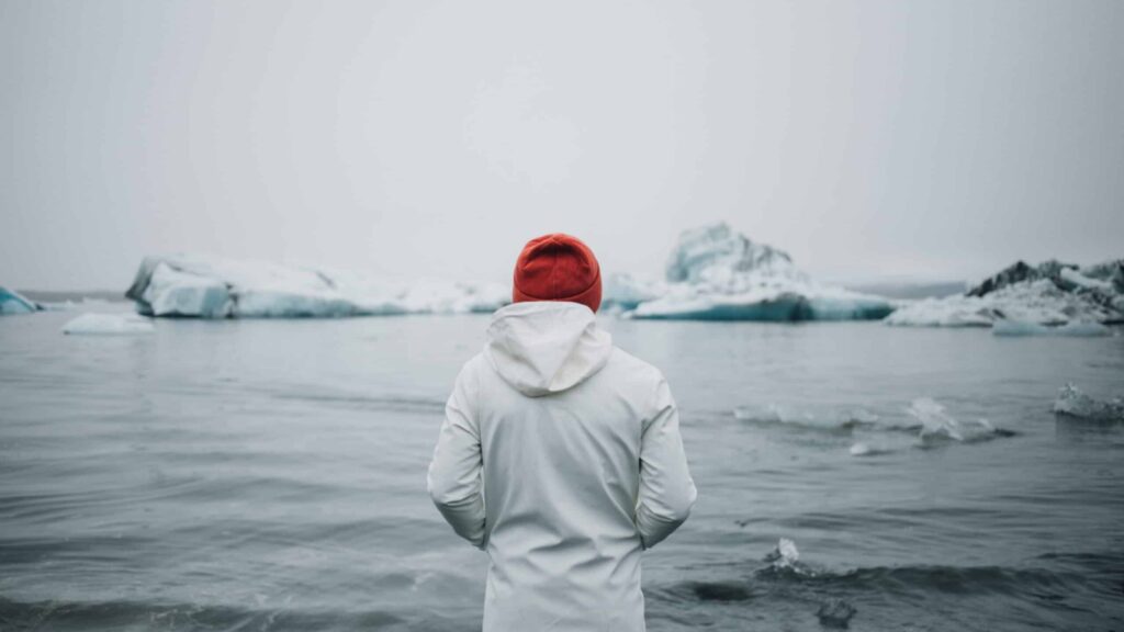 Tourist in a white jacket and red hat looking out to a glacier in water in iceland
