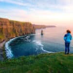 Woman standing on a grassy hilltop edge overlooking the ocean and vertical cliffs