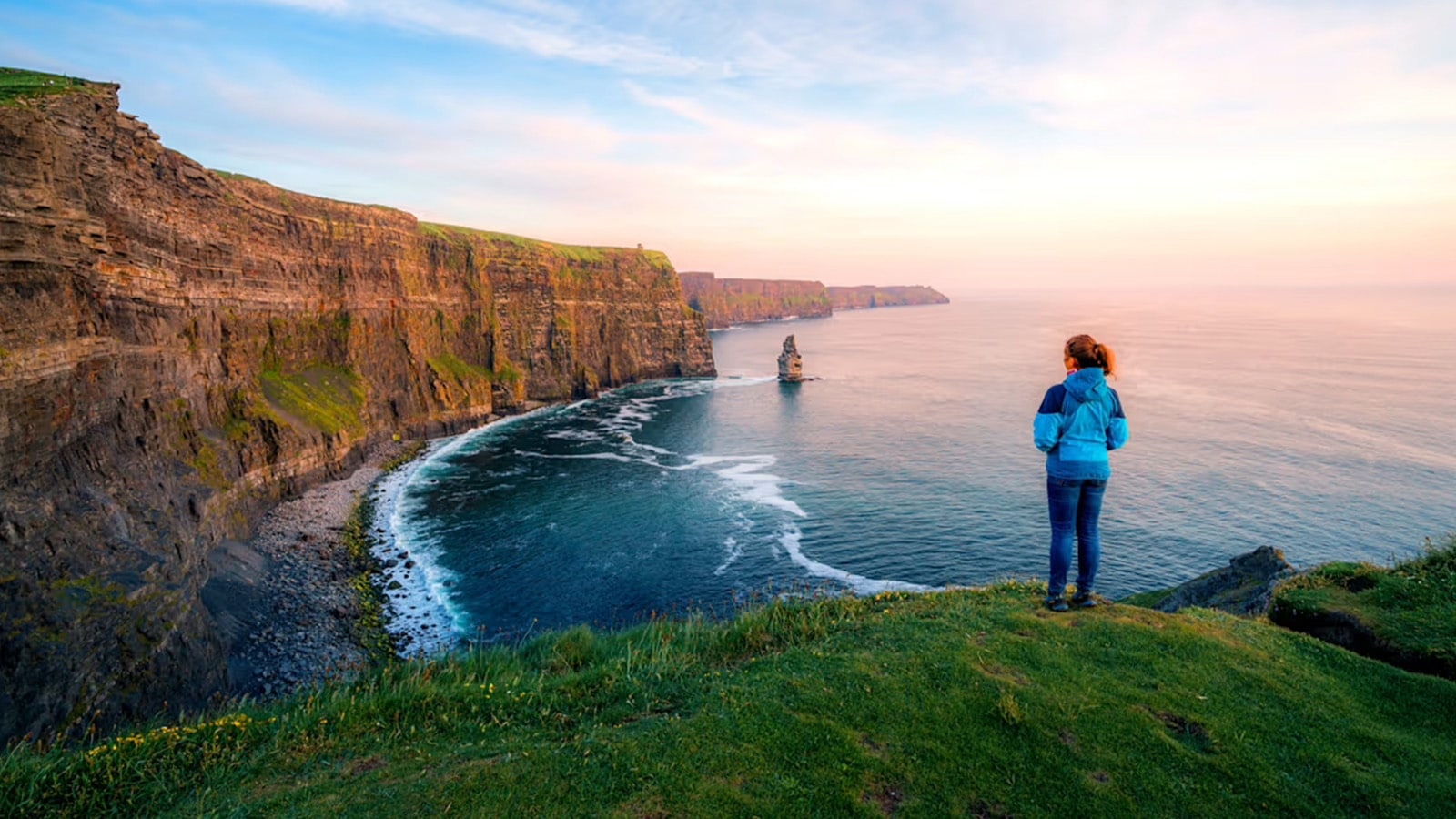 Woman standing on a grassy hilltop edge overlooking the ocean and vertical cliffs