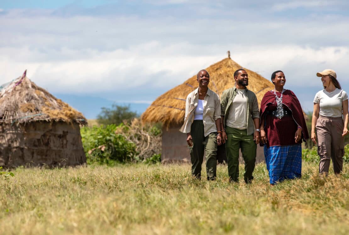 Four people walking through field with thatched-roof huts in the background