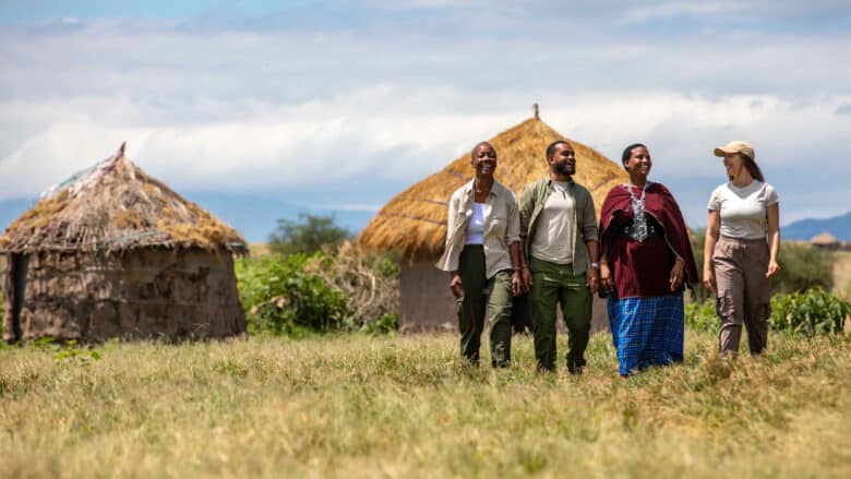 Four people walking through field with thatched-roof huts in the background