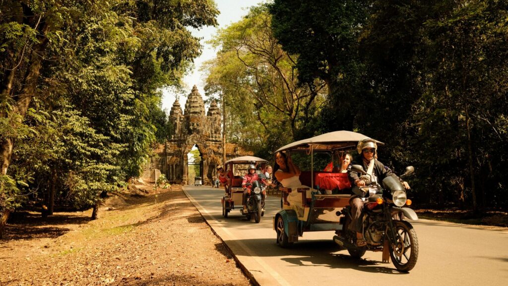 Two tuk tuks carrying travelers down a road by a temple in Cambodia on Coktiki's Ride Cambodia tour